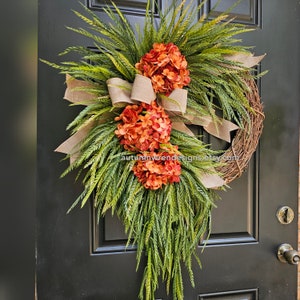 May include: A decorative wreath featuring orange hydrangeas and green foliage, accented with a burlap bow. The wreath is made of natural twigs and is designed for a front door. The text "autumnwrendesigns.etsy.com" is visible.