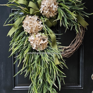May include: A decorative wreath featuring a natural grapevine base, adorned with faux greenery, beige hydrangea blooms, and a green ribbon. The wreath is designed for door or wall decor, adding a touch of nature to any space.