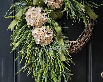 May include: A decorative wreath featuring a natural grapevine base, adorned with faux greenery, beige hydrangea blooms, and a green ribbon. The wreath is designed for door or wall decor, adding a touch of nature to any space.