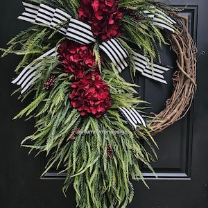 May include: A burgundy and green wreath with a black and white striped ribbon. The wreath is made of grapevine and has a cascading design. The wreath is decorated with burgundy hydrangeas and green sprigs.