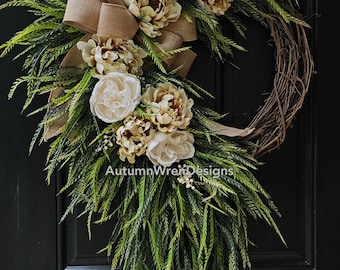 May include: A handcrafted grapevine wreath decorated with faux greenery, cream and tan peonies, and a burlap bow. The wreath is designed for a front door and features the text "AutumnWrenDesigns".