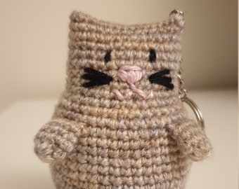 May include: A crocheted gray cat keychain with black eyes and a pink nose. The cat has a black embroidered mustache.