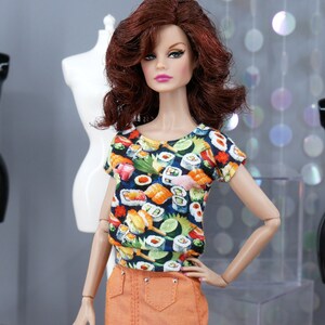 May include: A doll wearing a short-sleeved t-shirt with a colourful sushi print and an orange skirt. The doll has long, red hair and blue eyes.