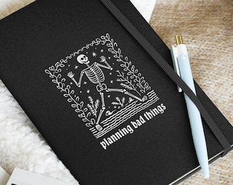 May include: Black notebook with a white illustration of a skeleton dancing in a floral frame. The text "planning bad things" is printed below the illustration.