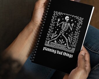 May include: A black spiral notebook with a white skeleton illustration and the text "planning bad things".