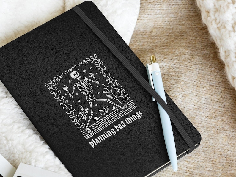 May include: Black notebook with a white illustration of a skeleton dancing in a floral frame. The text "planning bad things" is printed below the illustration.