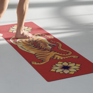 May include: A red yoga mat with a tiger illustration and a yellow flower design. The tiger is curled up with its head facing the viewer. The text "Strength & Grace" is printed on the mat.
