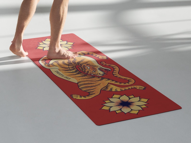 May include: A red yoga mat with a tiger illustration and a yellow flower design. The tiger is curled up with its head facing the viewer. The text "Strength & Grace" is printed on the mat.