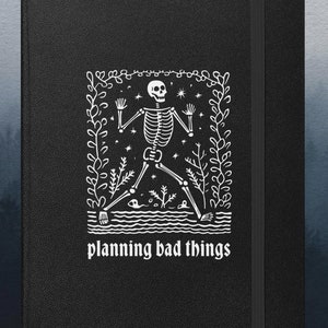May include: Black notebook with a white illustration of a skeleton in a forest setting. The text "planning bad things" is printed below the illustration.
