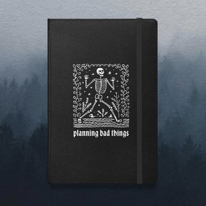 May include: Black notebook with a white illustration of a skeleton in a forest setting. The text "planning bad things" is printed below the illustration.