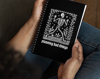 May include: Black spiral-bound notebook with a white illustration of a skeleton dancing in a floral frame. The text "planning bad things" is below the skeleton. The notebook is held in a person's hands.