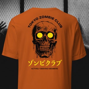 May include: Orange t-shirt with a black and yellow graphic of a skull with the text "Tokyo Zombie Club" and "ゾンビクラブ" (Zombie Club) and "Actively Seeking Members".
