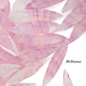 May include: Close-up of numerous, translucent, elongated, pink gemstones. The gemstones have a pointed shape and display subtle, parallel, lighter pink striations. The background is white, and the gemstones are scattered across the surface. The text "MrStones" is visible.