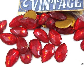 May include: A close-up of a pile of red, faceted, teardrop-shaped glass rhinestones. The rhinestones are scattered on a white surface. The rhinestones are vintage and have a slightly iridescent finish.