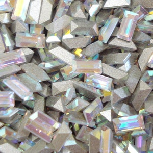 May include: A close-up image of a pile of iridescent, rectangular-shaped rhinestones. The rhinestones are scattered randomly and are various sizes. The rhinestones are a light, iridescent color with a rainbow sheen.