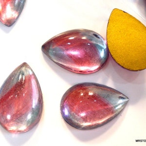 May include: Close-up of several iridescent teardrop-shaped beads. The beads have a mix of pink, purple, and silver hues. One bead is a solid yellow color. The beads are likely for jewelry making or craft projects.
