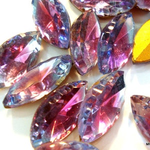 May include: A group of iridescent purple and blue glass rhinestones in a marquise shape. The rhinestones have a faceted design and are scattered on a white surface.