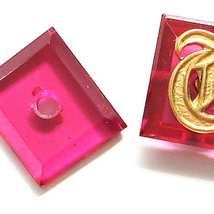 Op de afbeelding: Twee vierkante, roze glazen knopen met een gouden letter "T" op de ene knop. De andere knop heeft een gat in het midden.