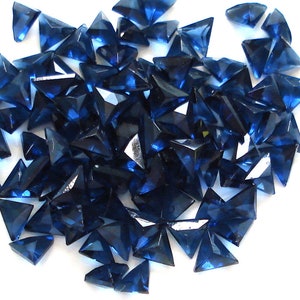 Puede incluir: Un montón grande de piedras de cristal triangulares de color azul oscuro. Las piedras de cristal son facetadas y tienen un acabado brillante.