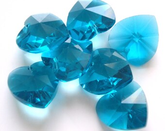 May include: A group of seven teal blue heart-shaped crystal beads. The beads are faceted and have a shiny finish.