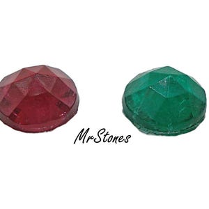 May include: Two faceted gemstones: one deep red and one emerald green. The gemstones are round with a domed top and a multi-faceted surface. The text "MrStones" is written in cursive between the stones.