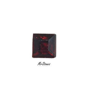 May include: A square-cut, dark red gemstone with a faceted surface. The gemstone is set against a white background. The text "Mr. Stones" is written in cursive below the gemstone.