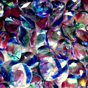 May include: A close-up of iridescent, faceted glass gems in shades of blue, purple, and green. The gems are scattered on a white surface.