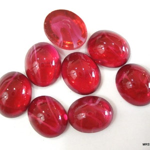 May include: A collection of eight oval-shaped, red glass cabochons. Each cabochon has a glossy finish with internal streaks of lighter red. The stones are arranged on a white surface, showcasing their vibrant color and reflective qualities.