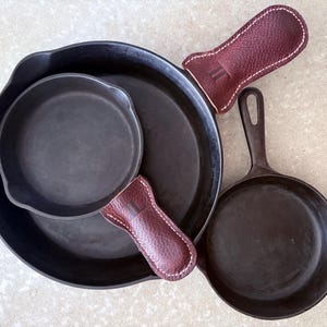 May include: A collection of black cast iron skillets of varying sizes, including a large skillet, a medium skillet, and a small skillet. Two burgundy leather handle covers with white stitching are also included.