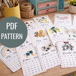 Puede incluir: Una colección de calendarios de 2026 con diseños de bordados florales para cada mes. Los calendarios están dispuestos sobre una superficie de madera, con el texto "PDF PATTERN" en un círculo. Cada calendario presenta una flor diferente.