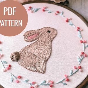 Puede incluir: Diseño de conejo bordado en un aro de madera. El conejo es de color marrón claro con el interior de las orejas rosado, rodeado por una corona floral con bayas rojas y rosas y hojas de color azul claro. El texto "PDF PATTERN" está en un círculo en la parte superior izquierda.