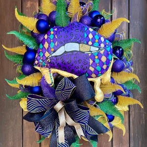 May include: A purple and gold Mardi Gras wreath with a large purple and gold lips design, a black and gold bow, and blue and gold ornaments.