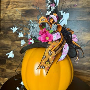 Puede incluir: Una calabaza naranja grande con un sombrero de bruja negro y una cinta rosa y naranja. El sombrero est&aacute; decorado con fantasmas blancos, gatos negros y una bruja. La calabaza est&aacute; iluminada con una cadena de luces.