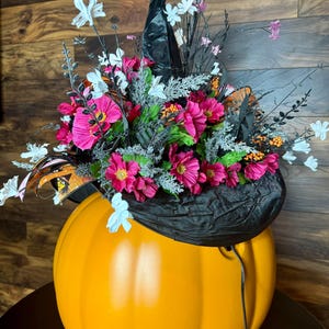Puede incluir: Una calabaza naranja grande con un sombrero de bruja negro decorado con flores rosas y blancas. El sombrero tiene una textura de terciopelo negro y est&aacute; adornado con estrellas brillantes y mariposas.