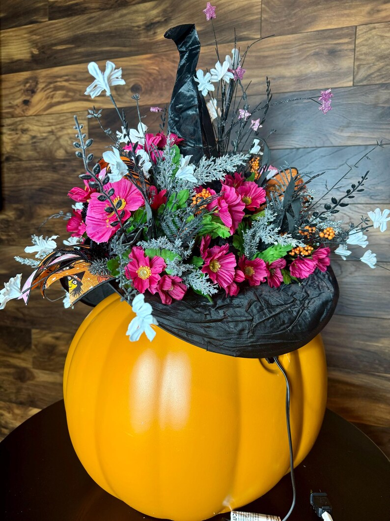 Puede incluir: Una calabaza naranja grande con un sombrero de bruja negro decorado con flores rosas y blancas. El sombrero tiene una textura de terciopelo negro y est&aacute; adornado con estrellas brillantes y mariposas.