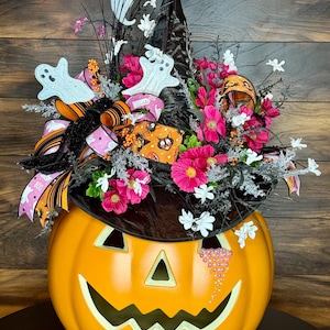 Puede incluir: Una calabaza naranja grande con una cara de calabaza tallada est&aacute; decorada con un sombrero de bruja negro, flores rosas y blancas y fantasmas blancos. La calabaza tiene purpurina rosa en el lateral.
