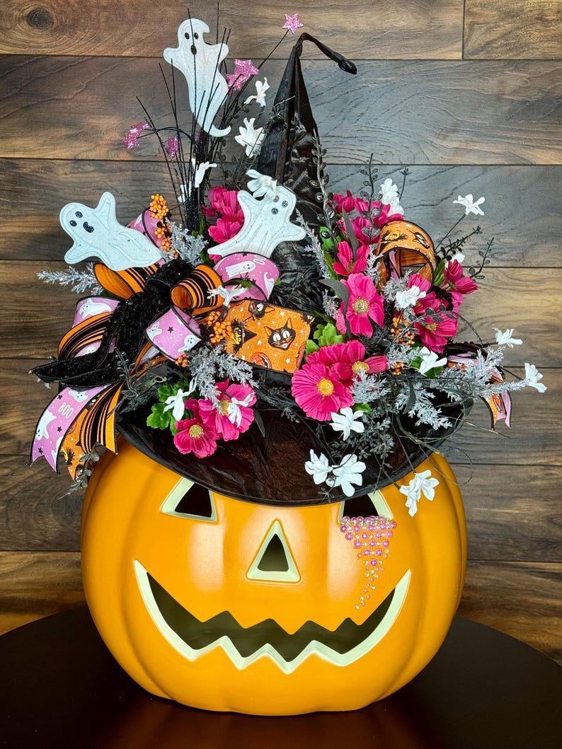 Puede incluir: Una calabaza naranja grande con una cara de calabaza tallada est&aacute; decorada con un sombrero de bruja negro, flores rosas y blancas y fantasmas blancos. La calabaza tiene purpurina rosa en el lateral.