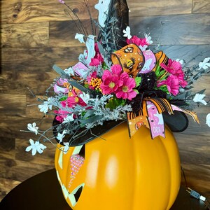 Puede incluir: Una calabaza naranja grande con un sombrero de bruja en la parte superior. El sombrero es negro con una cinta rosa y negra y un fantasma blanco. La calabaza tiene una cara de jack-o'-lantern con diamantes de imitaci&oacute;n rosas.