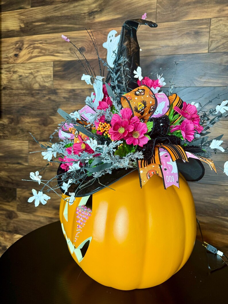 Puede incluir: Una calabaza naranja grande con un sombrero de bruja en la parte superior. El sombrero es negro con una cinta rosa y negra y un fantasma blanco. La calabaza tiene una cara de jack-o'-lantern con diamantes de imitaci&oacute;n rosas.