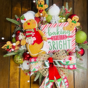 May include: A festive Christmas wreath with a gingerbread theme. It features a large gingerbread man, decorative ornaments, and a sign that reads "baking spirits bright". The wreath is adorned with red, green, and white accents, and a large bow.
