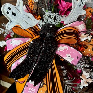 Puede incluir: Un centro de mesa tem&aacute;tico de Halloween con una cinta a rayas negra y naranja con purpurina negra, una cinta rosa con fantasmas blancos y estrellas, y la palabra "BOO" impresa en ella. El centro de mesa tambi&eacute;n incluye recortes de fantasmas blancos, acentos florales en blanco y negro y bayas naranjas y rosas.