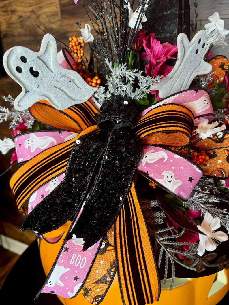Puede incluir: Un centro de mesa tem&aacute;tico de Halloween con una cinta a rayas negra y naranja con purpurina negra, una cinta rosa con fantasmas blancos y estrellas, y la palabra "BOO" impresa en ella. El centro de mesa tambi&eacute;n incluye recortes de fantasmas blancos, acentos florales en blanco y negro y bayas naranjas y rosas.