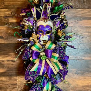 May include: A Mardi Gras themed wreath featuring a decorative mask with purple, gold, and green accents. The wreath includes ribbons, bows, and floral elements in matching colors, creating a festive and eye-catching design.