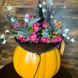 Puede incluir: Una calabaza naranja grande con un sombrero de bruja negro en la parte superior. El sombrero est&aacute; decorado con flores rosas y ramitas blancas. La calabaza est&aacute; iluminada con una cadena de luces.