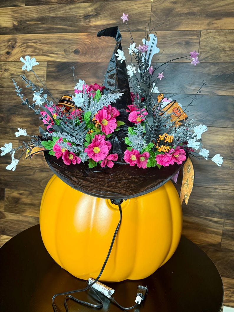 Puede incluir: Una calabaza naranja grande con un sombrero de bruja negro en la parte superior. El sombrero est&aacute; decorado con flores rosas y ramitas blancas. La calabaza est&aacute; iluminada con una cadena de luces.