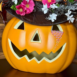 Puede incluir: Calabaza naranja de Halloween con un sombrero de bruja negro decorado con flores rosas y blancas. La calabaza tiene una cara tallada con una l&aacute;grima hecha de diamantes de imitaci&oacute;n rosas.