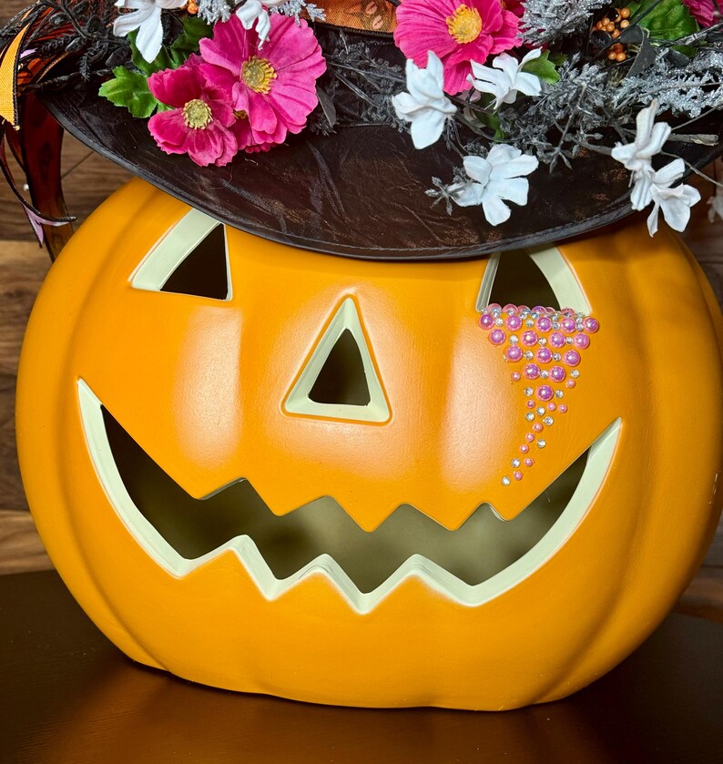 Puede incluir: Calabaza naranja de Halloween con un sombrero de bruja negro decorado con flores rosas y blancas. La calabaza tiene una cara tallada con una l&aacute;grima hecha de diamantes de imitaci&oacute;n rosas.