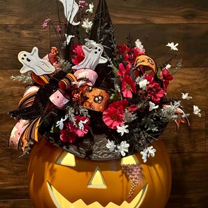 Puede incluir: Una calabaza iluminada con forma de cara de jack-o'-lantern con un sombrero de bruja negro decorado con flores rosas y blancas, una cinta negra y naranja y fantasmas blancos.