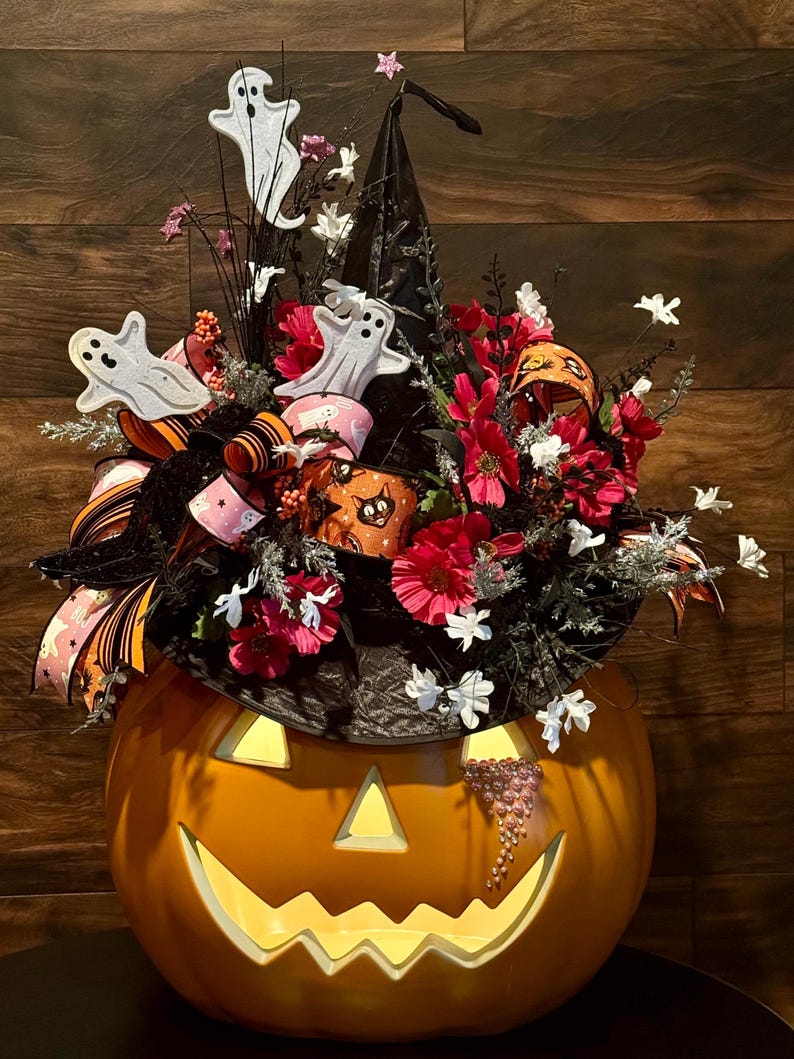 Puede incluir: Una calabaza iluminada con forma de cara de jack-o'-lantern con un sombrero de bruja negro decorado con flores rosas y blancas, una cinta negra y naranja y fantasmas blancos.
