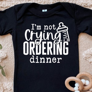 May include: Black baby onesie with white text that reads "I'm not crying I'm ordering dinner" and a graphic of a baby bottle.